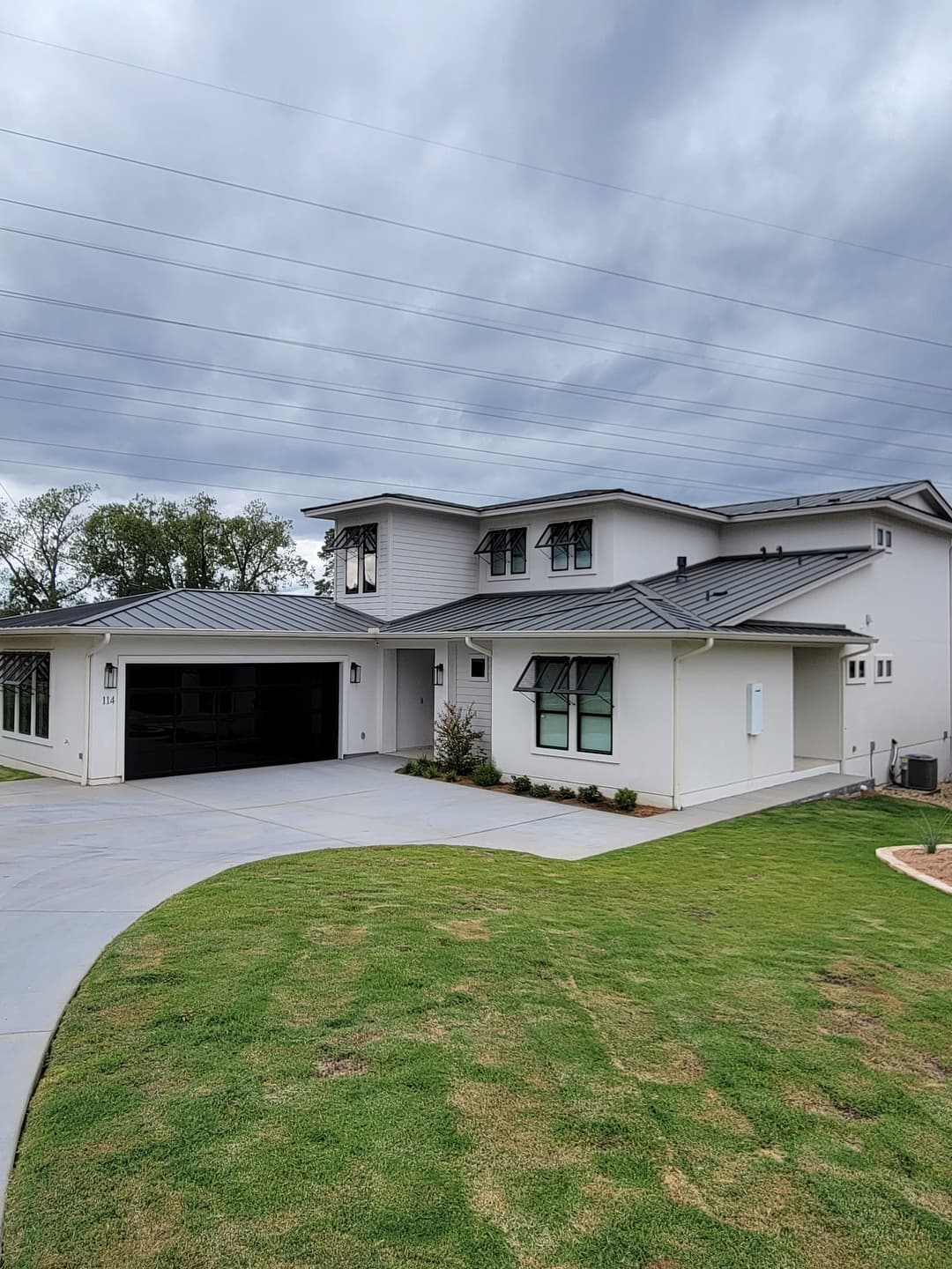 Modern white house with gray roof, landscaped yard, and driveway under cloudy sky.