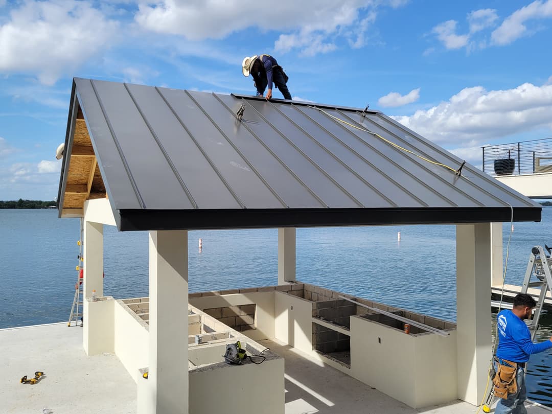 Construction worker installing a metal roof on a waterfront pavilion under a blue sky.