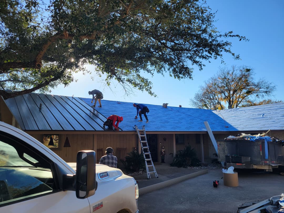 Workers installing a metal roof on a house with trees and a pickup truck nearby.