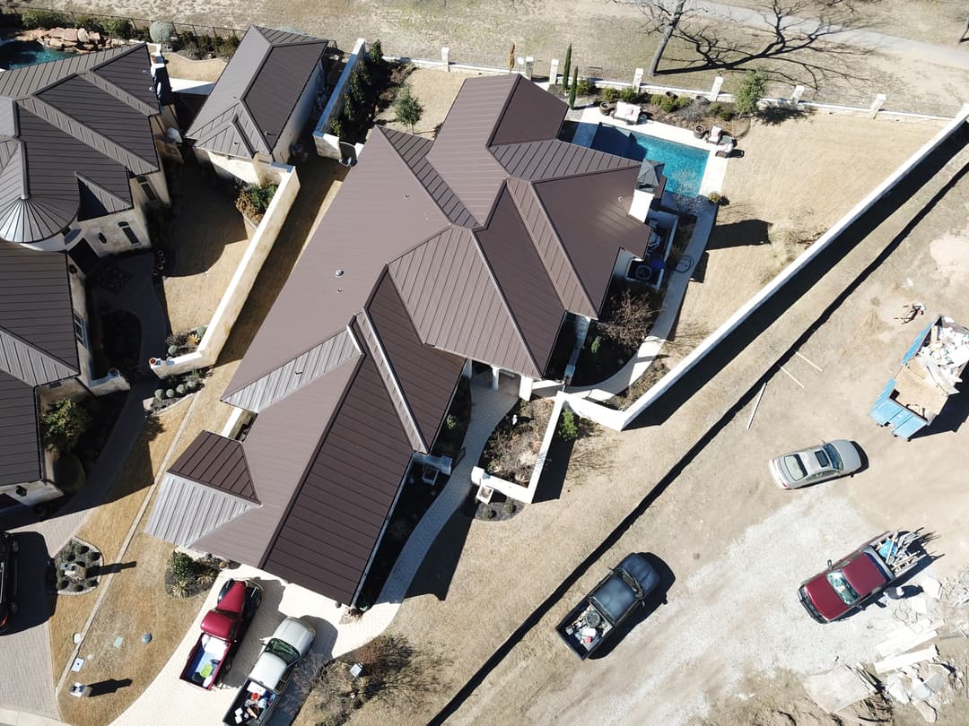 Aerial view of a modern home with a brown roof, surrounded by a landscaped yard and parked cars.