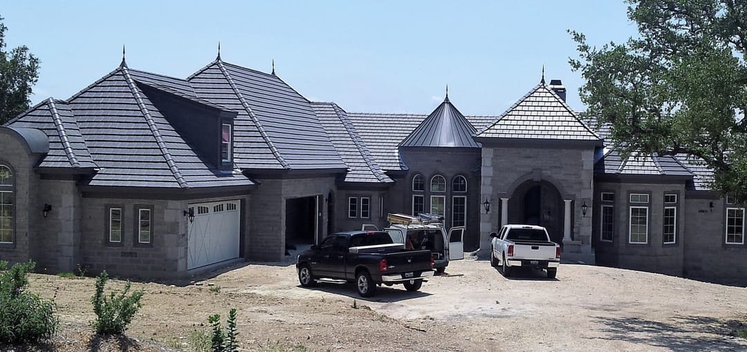 Modern stone house with multiple peaked roofs and two parked trucks in a gravel driveway.