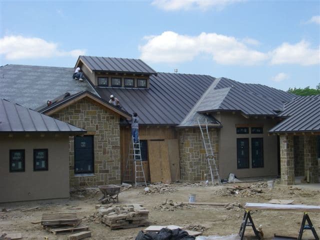 Construction workers installing a metal roof on a modern stone home under a blue sky.