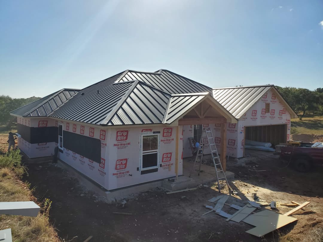 Newly constructed house with a metal roof, surrounded by construction materials and workers.