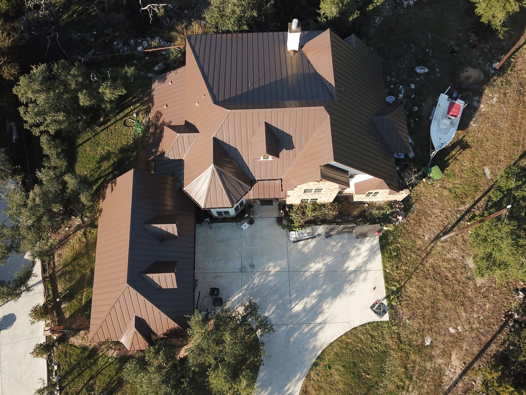 Aerial view of a large house with a brown metal roof, surrounded by greenery and a boat nearby.