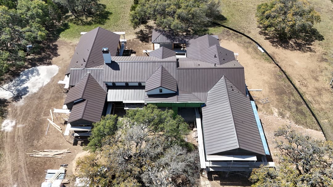 Aerial view of a large, modern home under construction with a dark metal roof and surrounding trees.